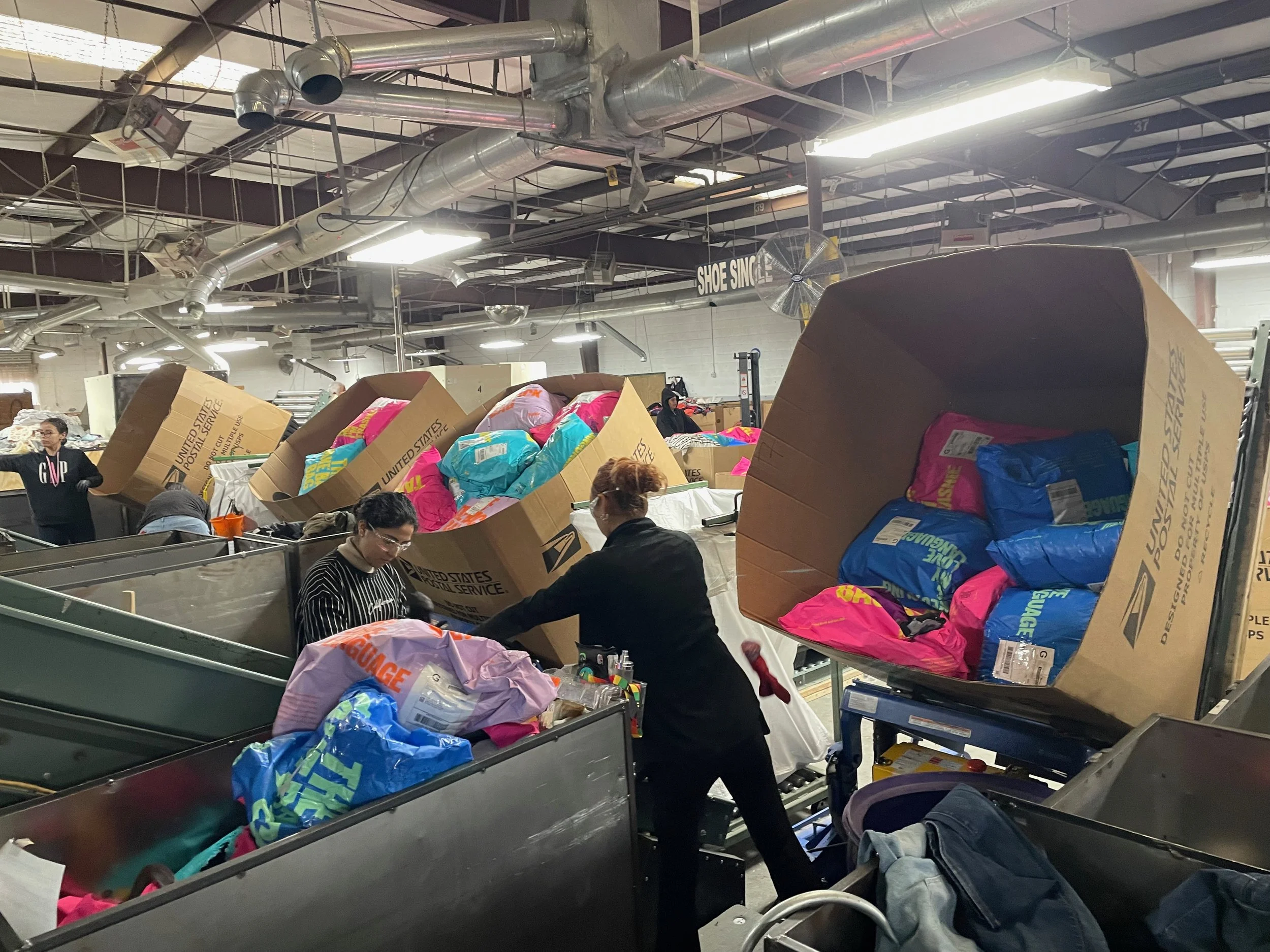 A warehouse filled with large boxes holding pink and blue plastic bags with mailers attached. Three women are processing the bags.