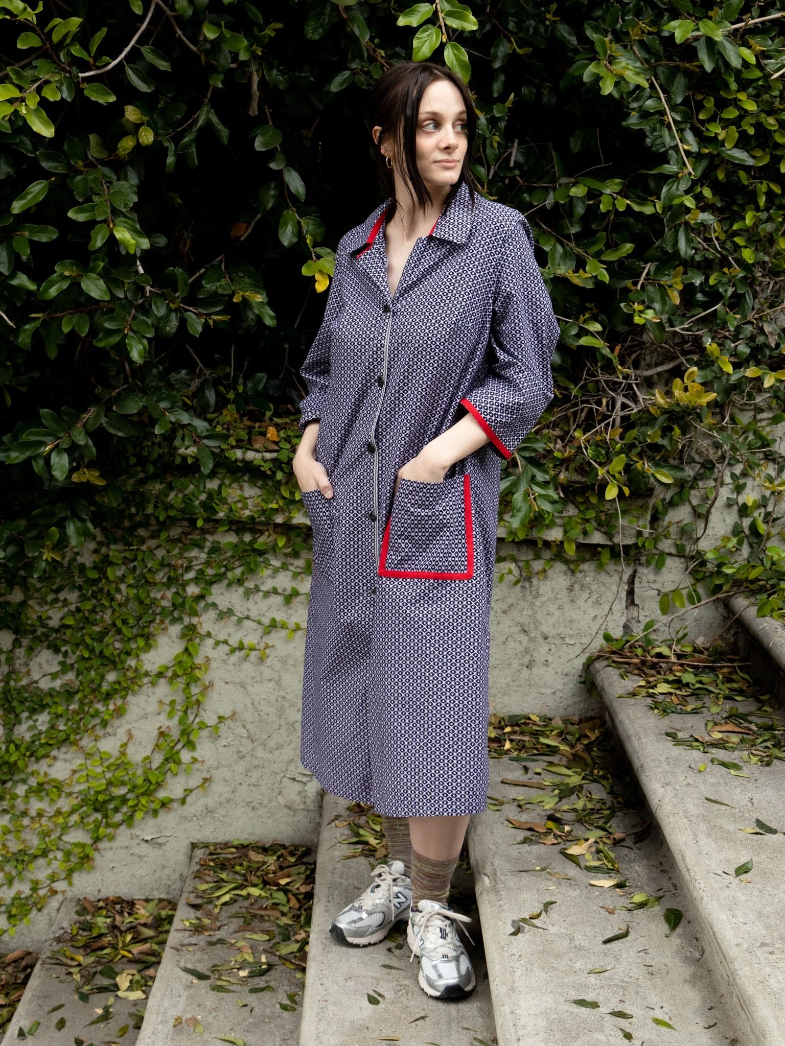 A white woman with dark hair is wearing a long blue jacket with red lined pockets and sneakers.