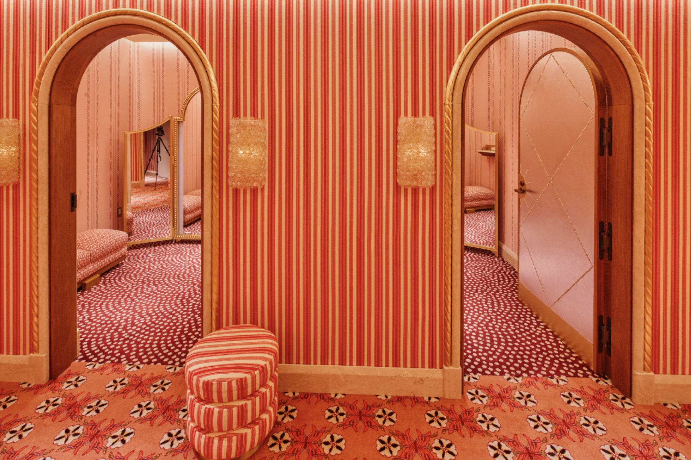 A luxurious retail fitting area featuring rich coral and red striped wallpaper, arched doorways with rope detailing, and coordinating patterned carpeting.