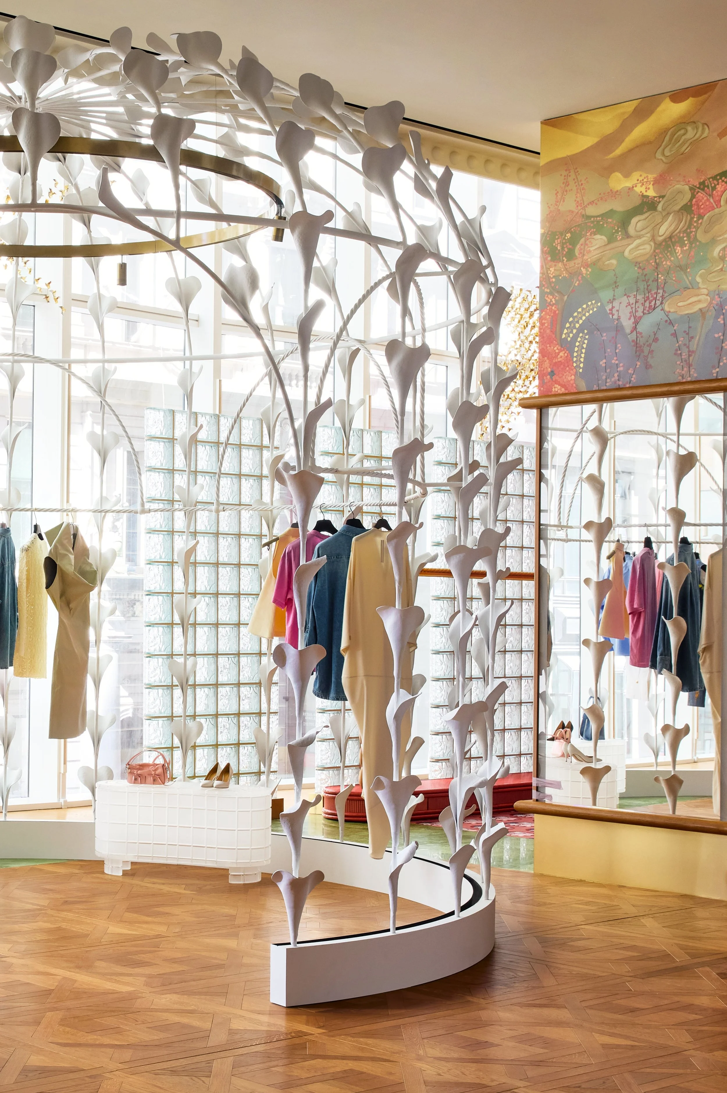An elegant fashion retail space with a striking white sculptural tree installation as the centerpiece, surrounded by clothing displays on brass fixtures.
