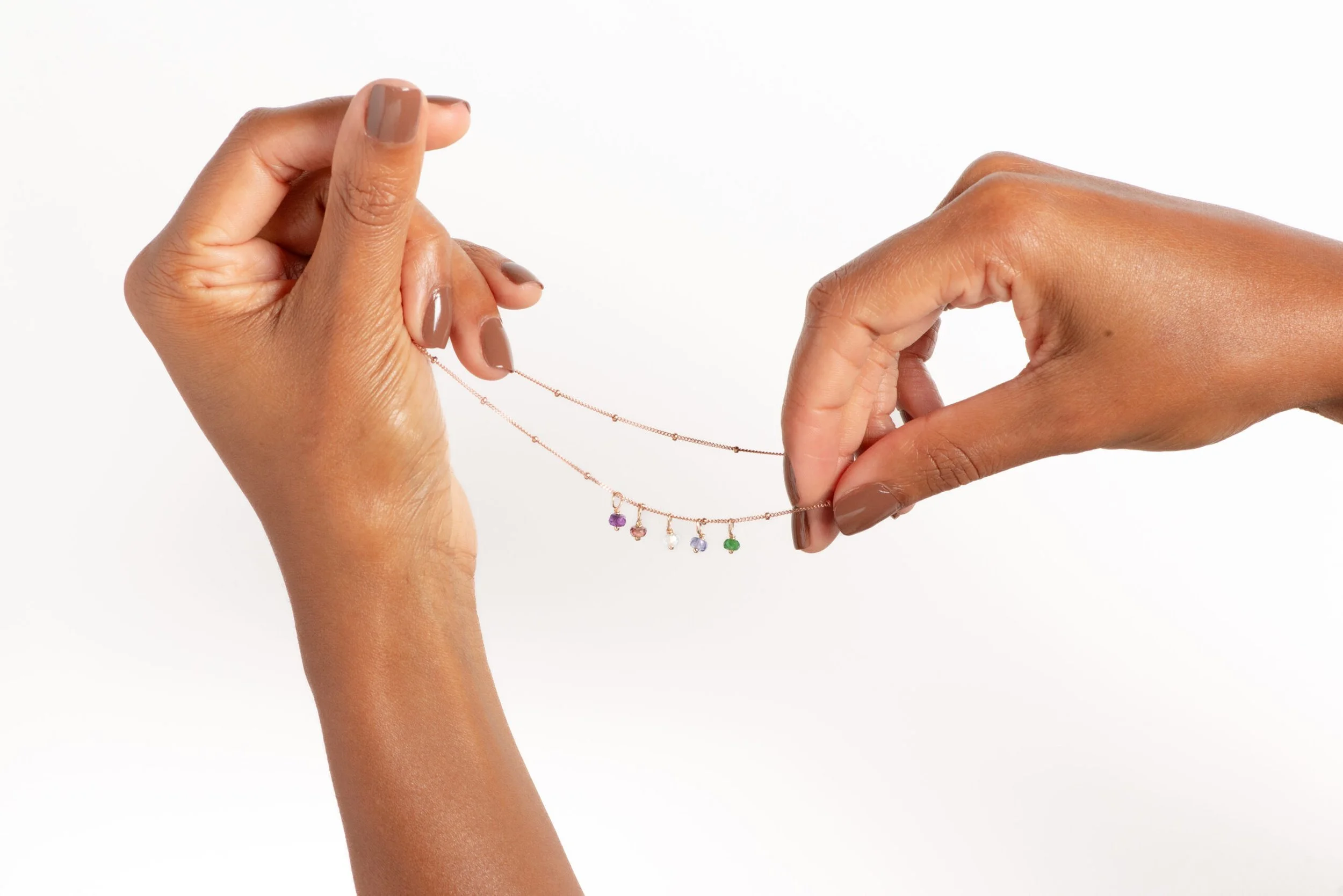 Two hands are holding a gold necklace with colorful stones hanging off of it with a white background.