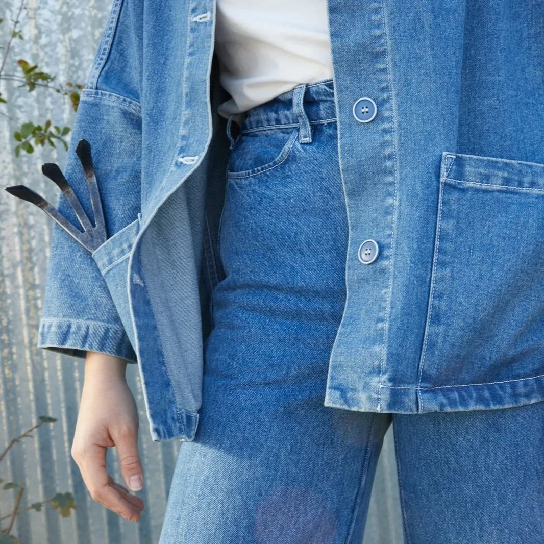 A woman is wearing blue jeans and a blue jean jacket with a garden tool in her pocket.