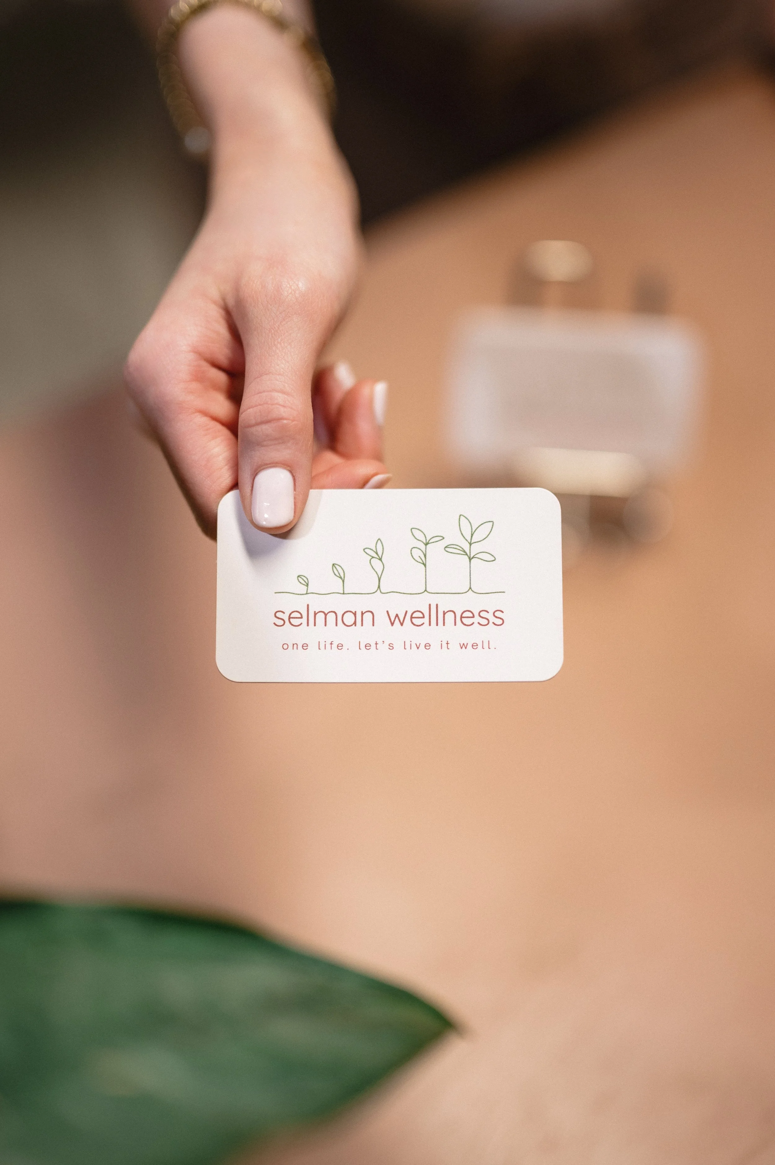 A person holding a business card for Selman Wellness, which features a minimalist illustration of plant sprouts and the tagline 'one life. let's live it well.'