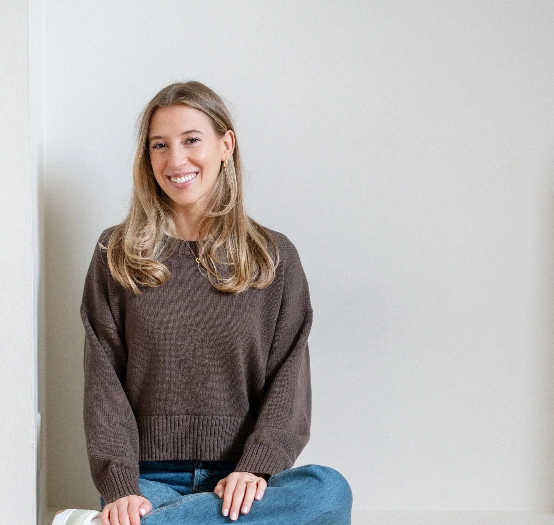 A smiling young woman with long blonde hair, wearing a brown sweater and blue jeans, sitting on the floor against a plain white wall.