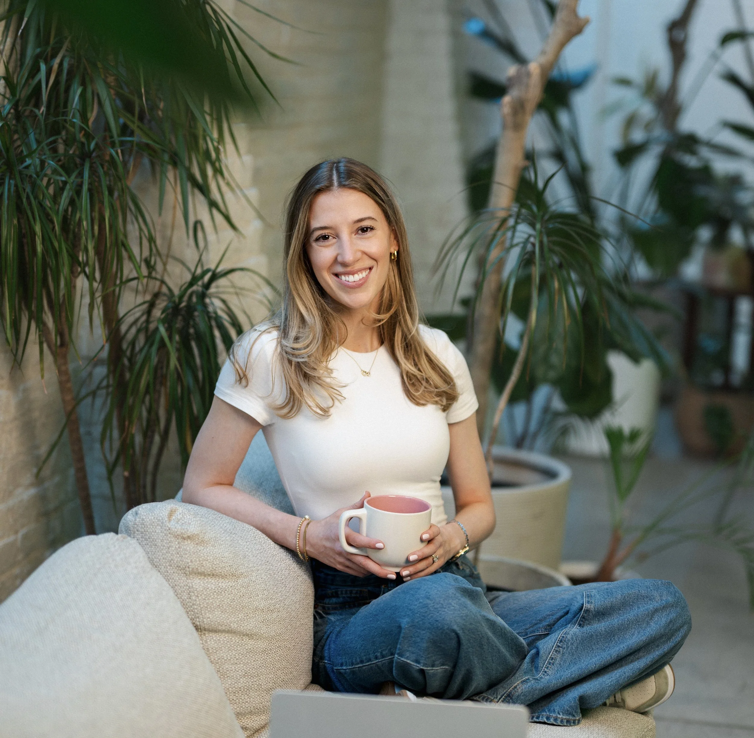 A woman sitting on a beige sofa, smiling, holding a white mug with a pink interior. She is wearing a white t-shirt and jewelry. There are large green plants behind her in an cozy setting.