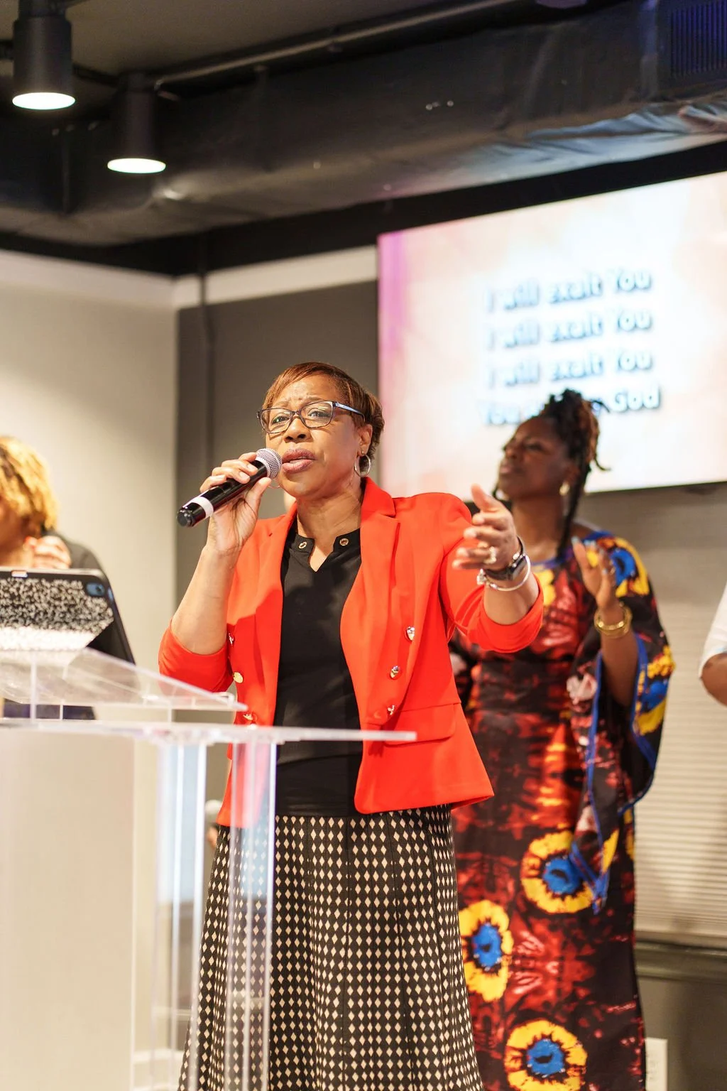 A woman in a red blazer and black blouse holding a microphone, Leading worship passionately, She stands in front of a screen displaying lyrics or words.