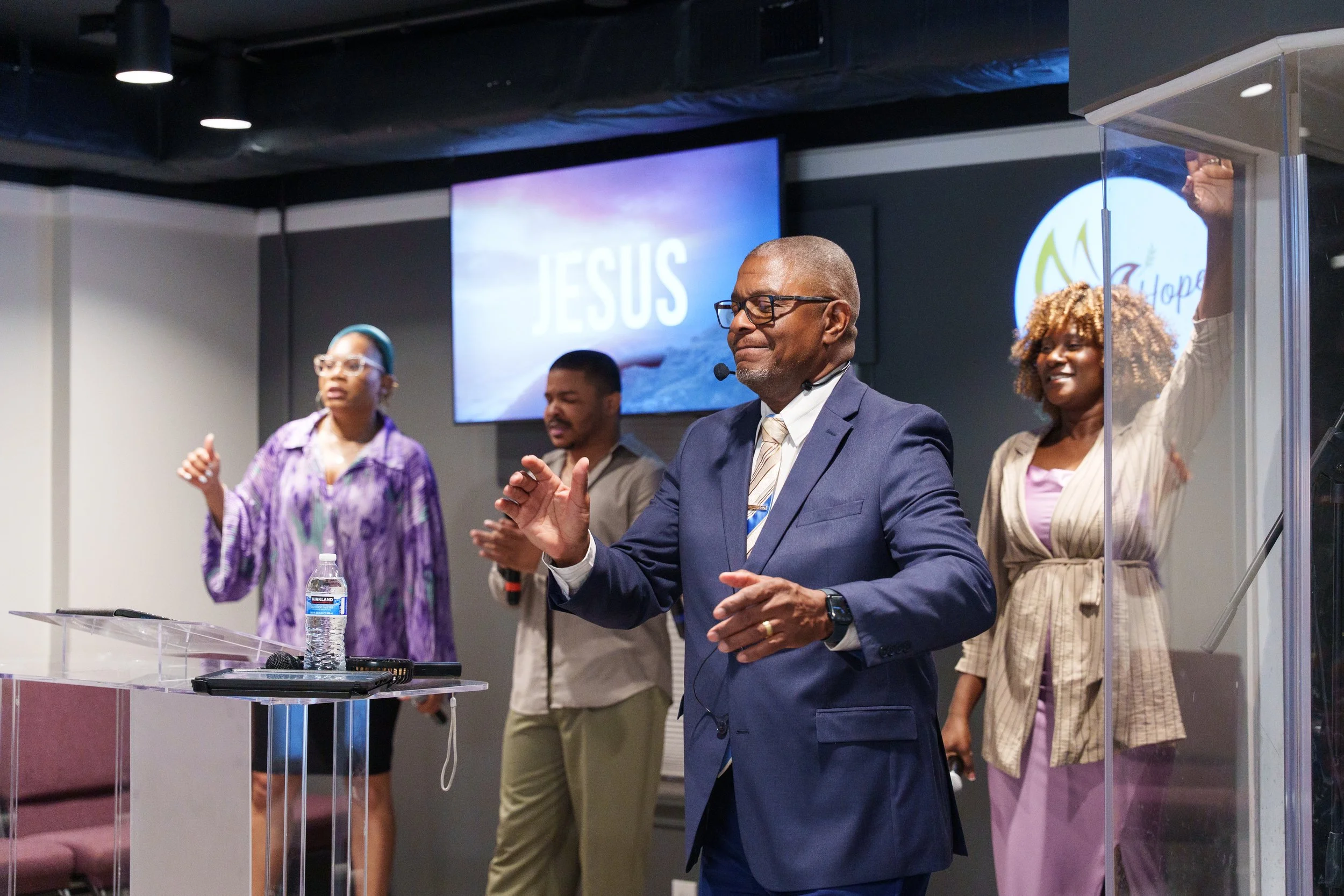 A group of four diverse people praying or worshiping in a church during a service, with the word 'JESUS' on a screen in the background.