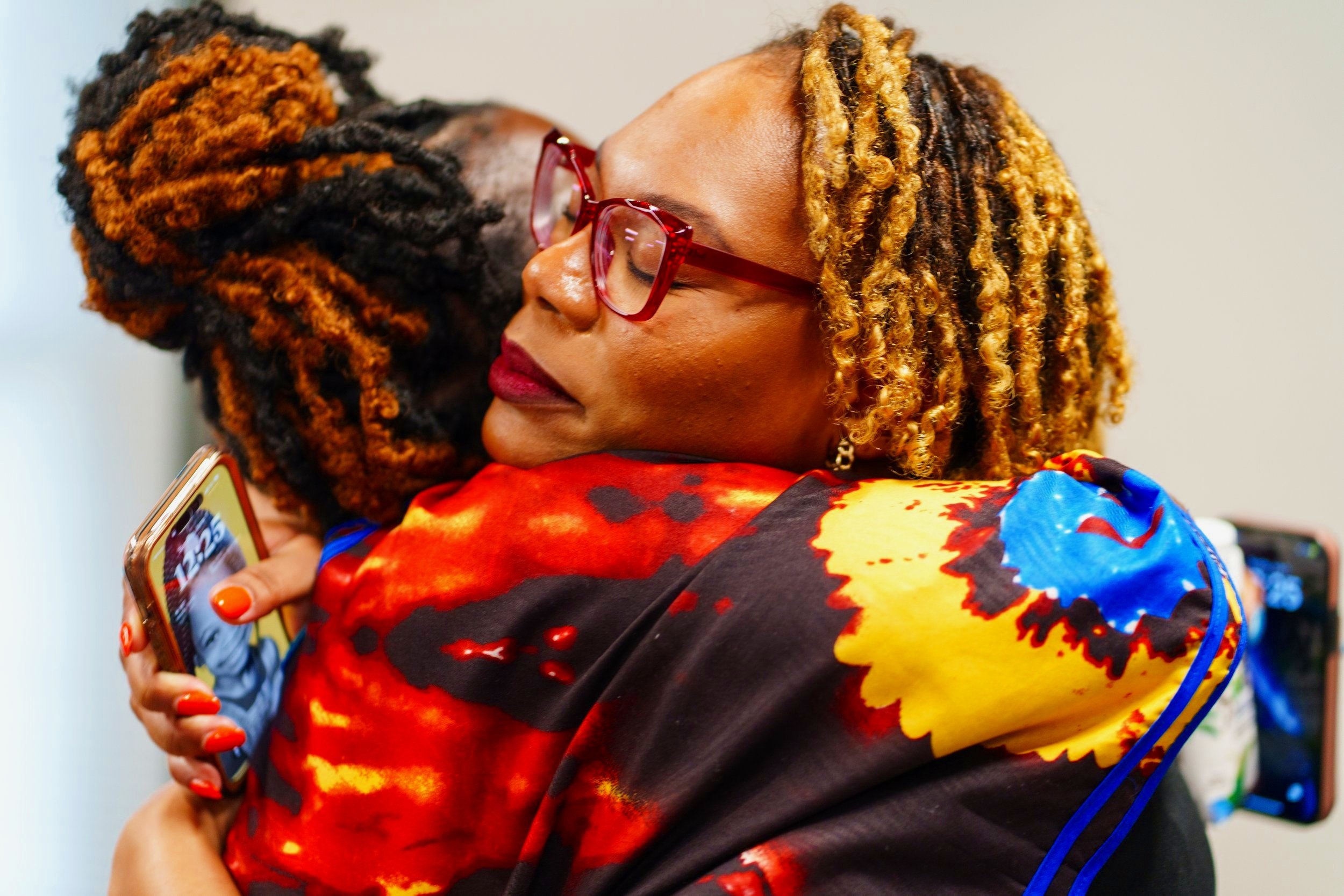 Two women embracing each other warmly; one woman with red glasses, colorful patterned jacket, and blonde curly hair; the other woman with dreadlocked hair with orange tips.