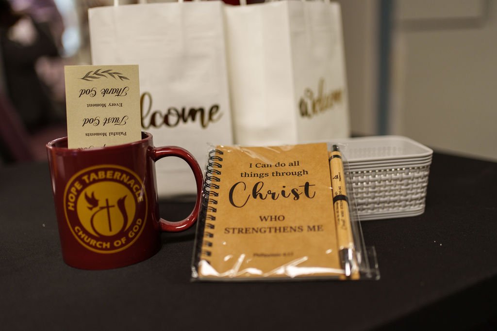 A red mug with Hope Tabernacle Church of God logo, a Bible verse card, a yellow pen, and a white basket on a table with white bags in the background.