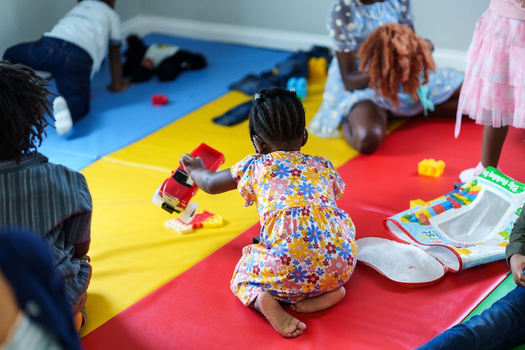 Children playing on colorful mats, with a girl in a floral dress kneeling and holding a toy fire truck.