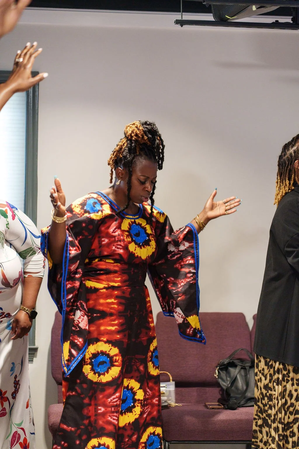 A woman with her eyes closed and hands raised, wearing a colorful dress with bold floral patterns, standing in a prayer or worship pose.