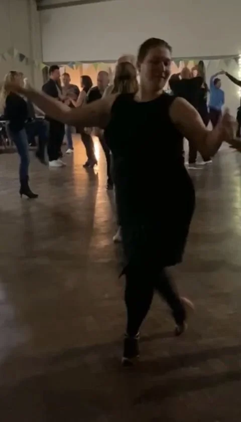 Woman dancing in a black dress at a social event with other people dancing in the background.