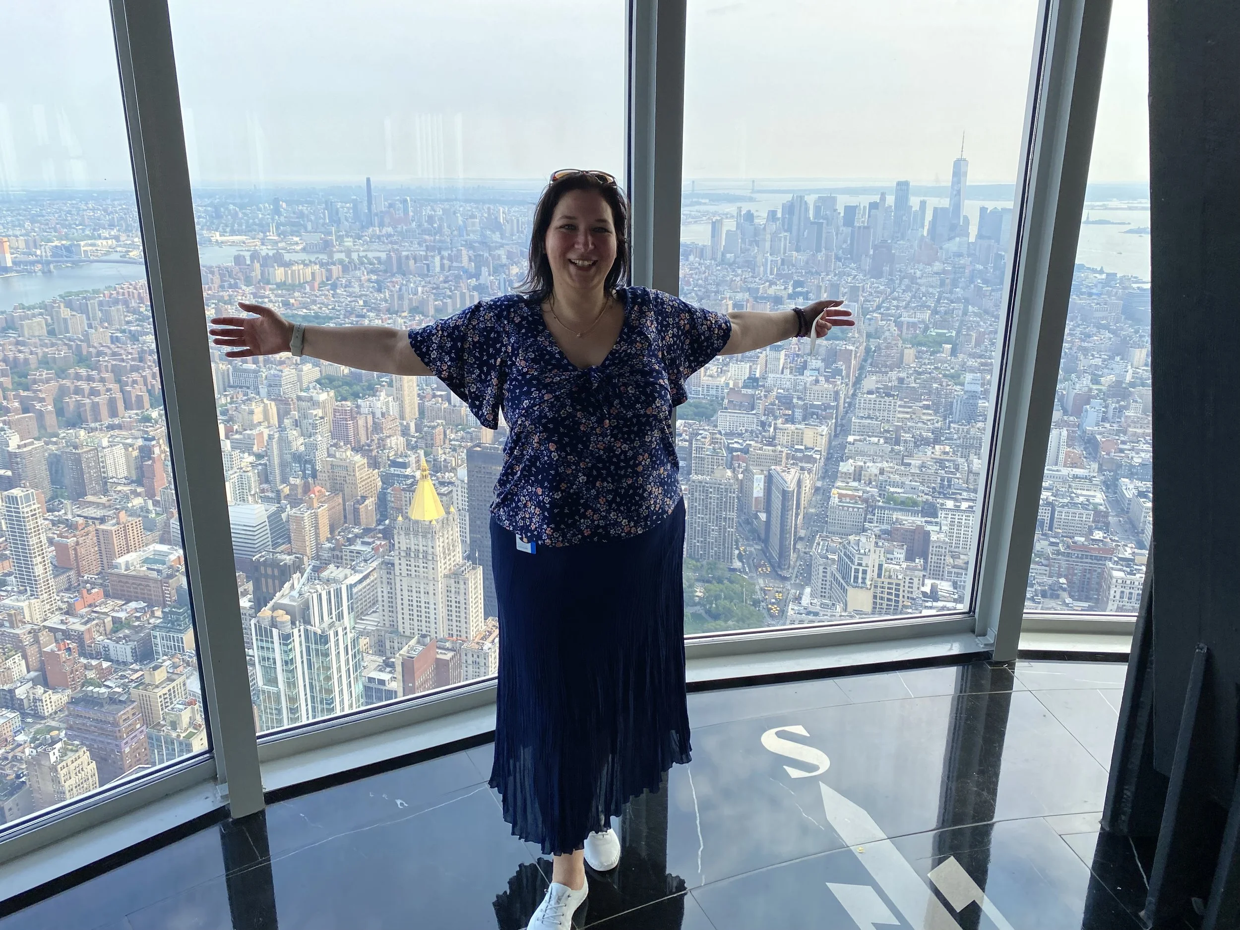Woman standing with arms outstretched in front of the Empire State Building's large window with New York's city skyline view, including skyscrapers and the Hudson River, in the background.