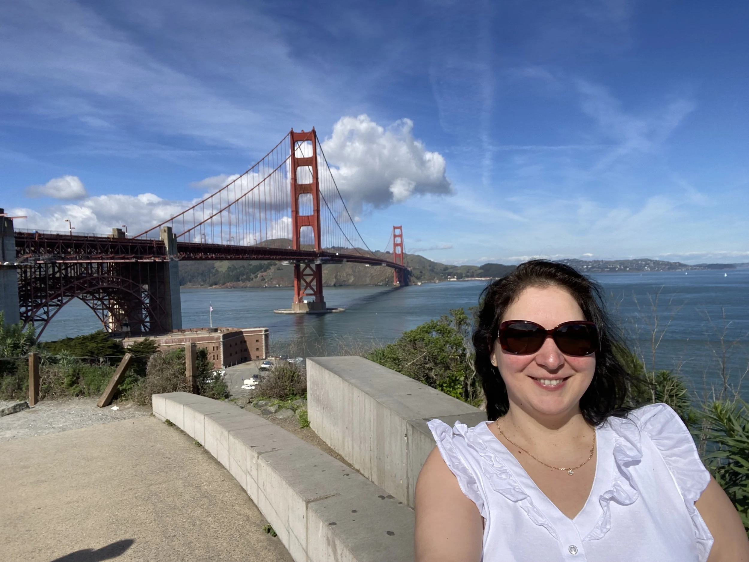 A woman smiling in front of the Golden Gate Bridge during the daytime, wearing sunglasses and a white top.