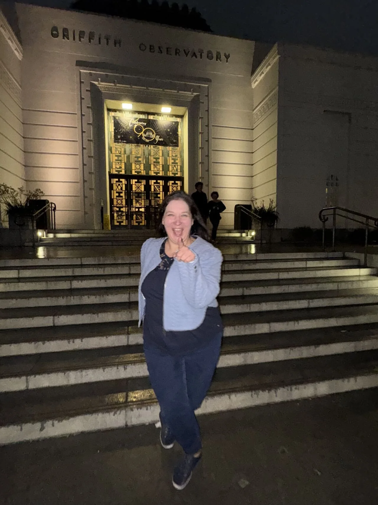 A woman smiling widely and pointing at the camera while standing on steps in front of the Griffith Observatory by Hollywood at night.