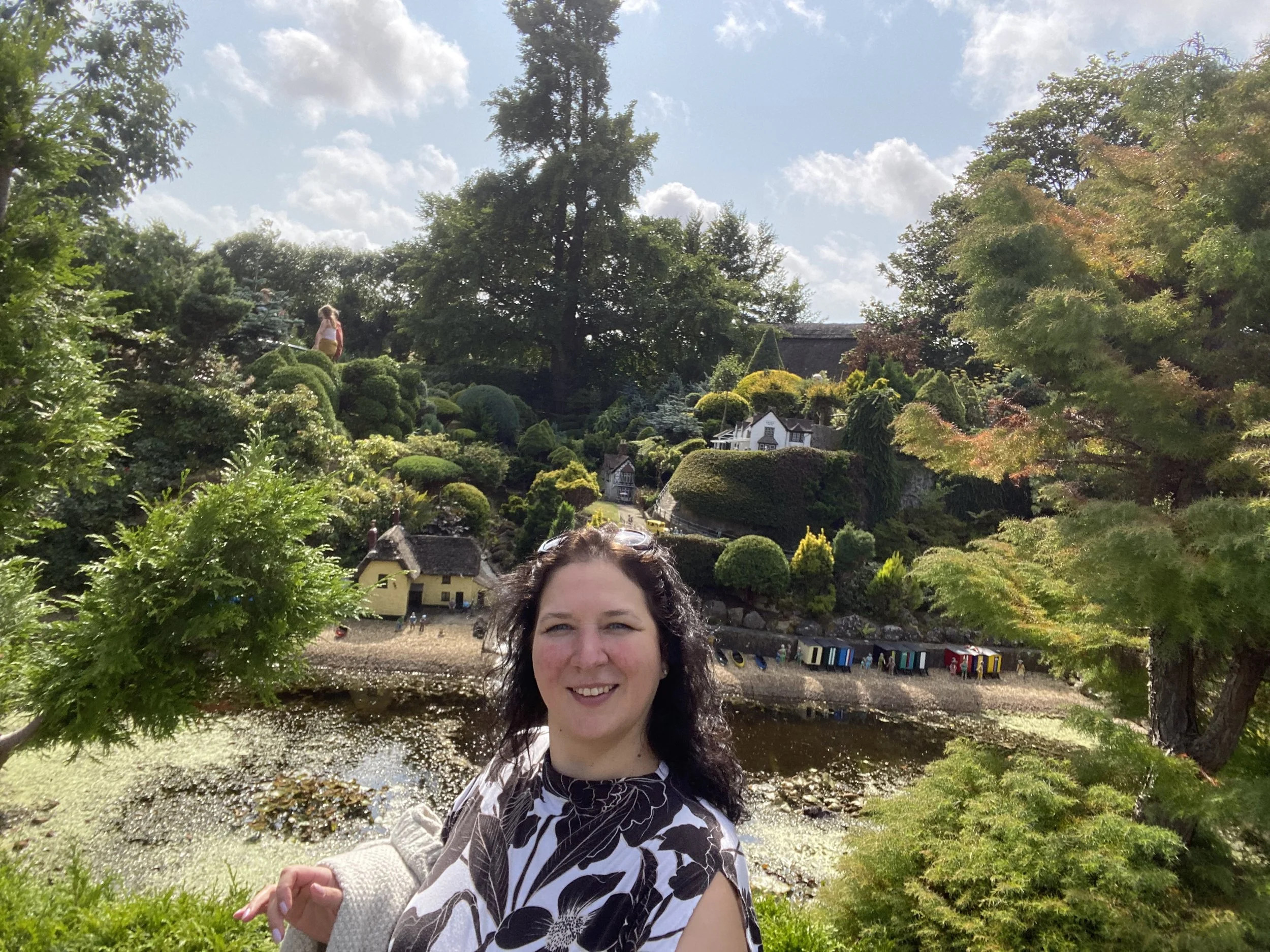 Woman smiling in a garden with miniature houses and lush green trees in the background.