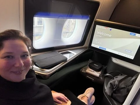A woman sitting at a first-class private seat in an airplane seat by the window, with a personal entertainment screen displaying a welcome message.