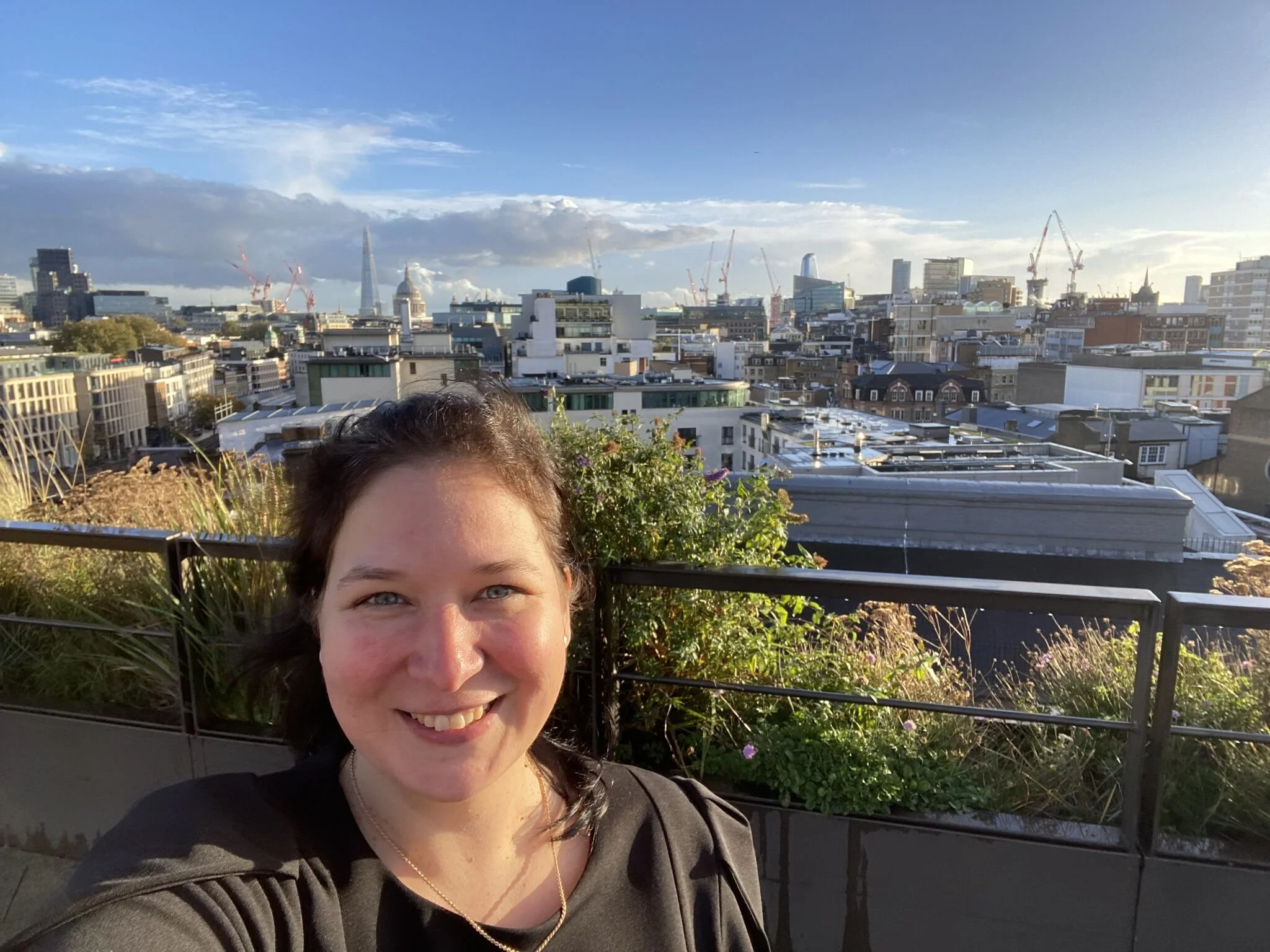 A woman taking a selfie on a rooftop with London city skyline and blue sky in the background.
