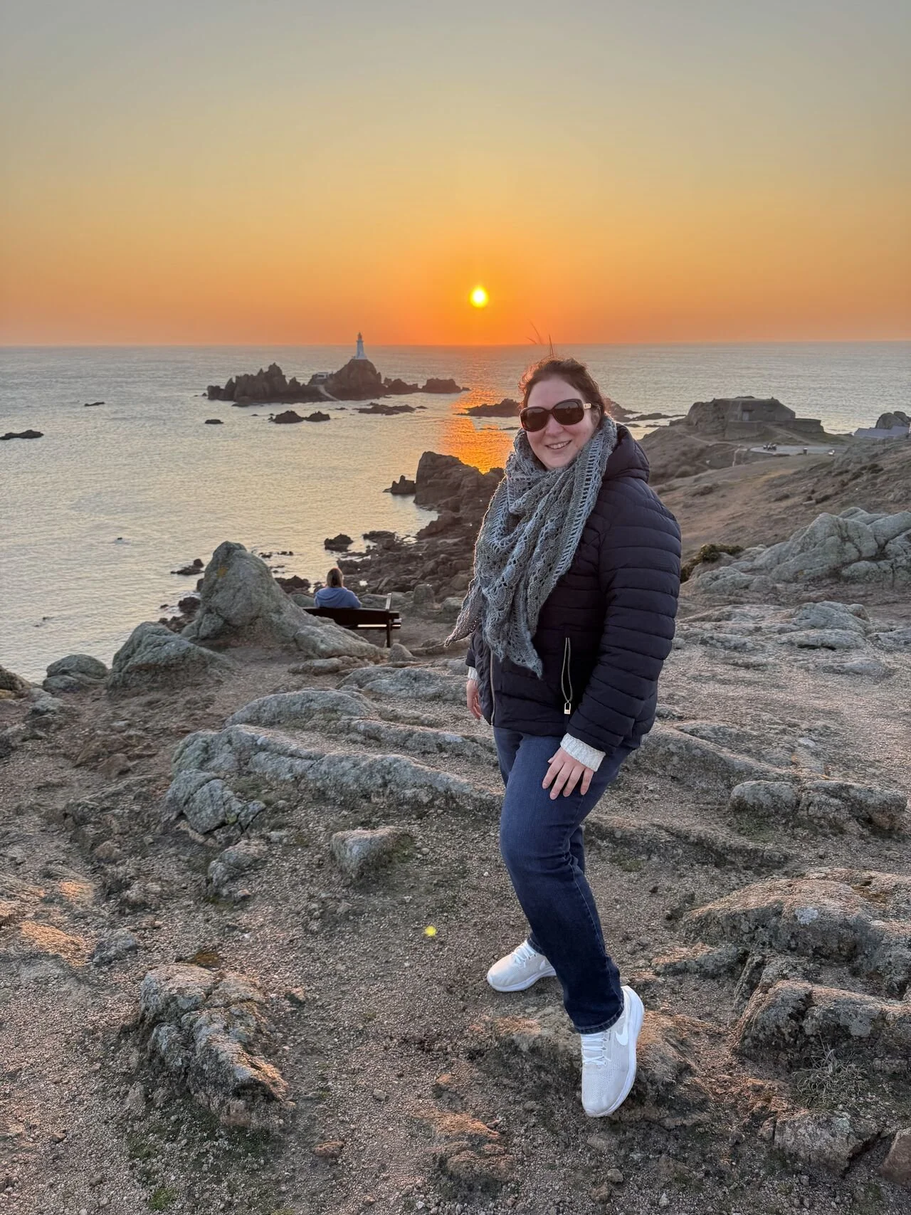 A woman with dark hair, wearing sunglasses, a green scarf, a navy jacket, jeans, and beige Nike sneakers, stands on rocky terrain near the coast with a sunset over the ocean with a lighthouse in the background.