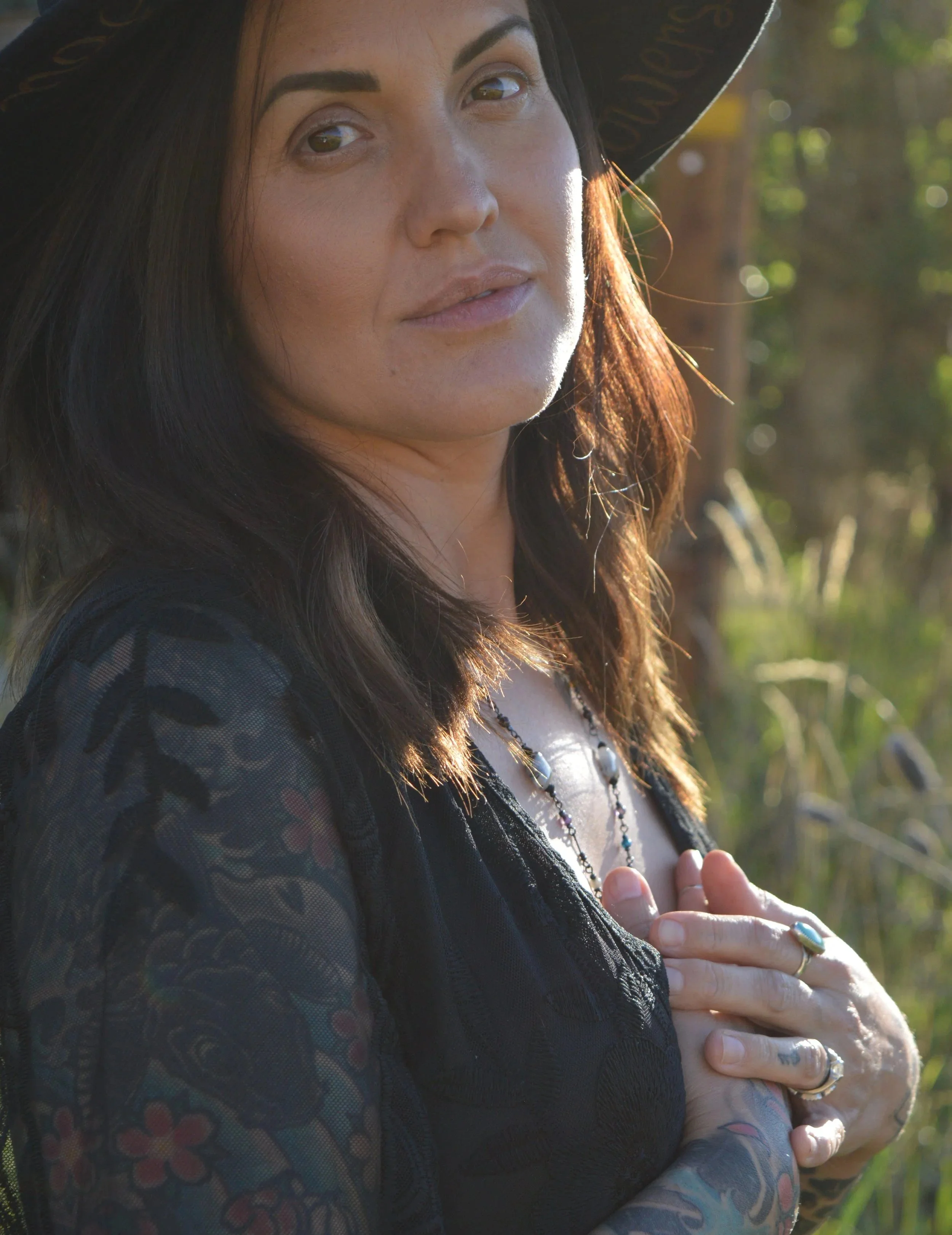 A woman with dark hair wearing a black hat and a black lace top, standing outdoors in a wooded area, looking to the side with sunlight on her face.