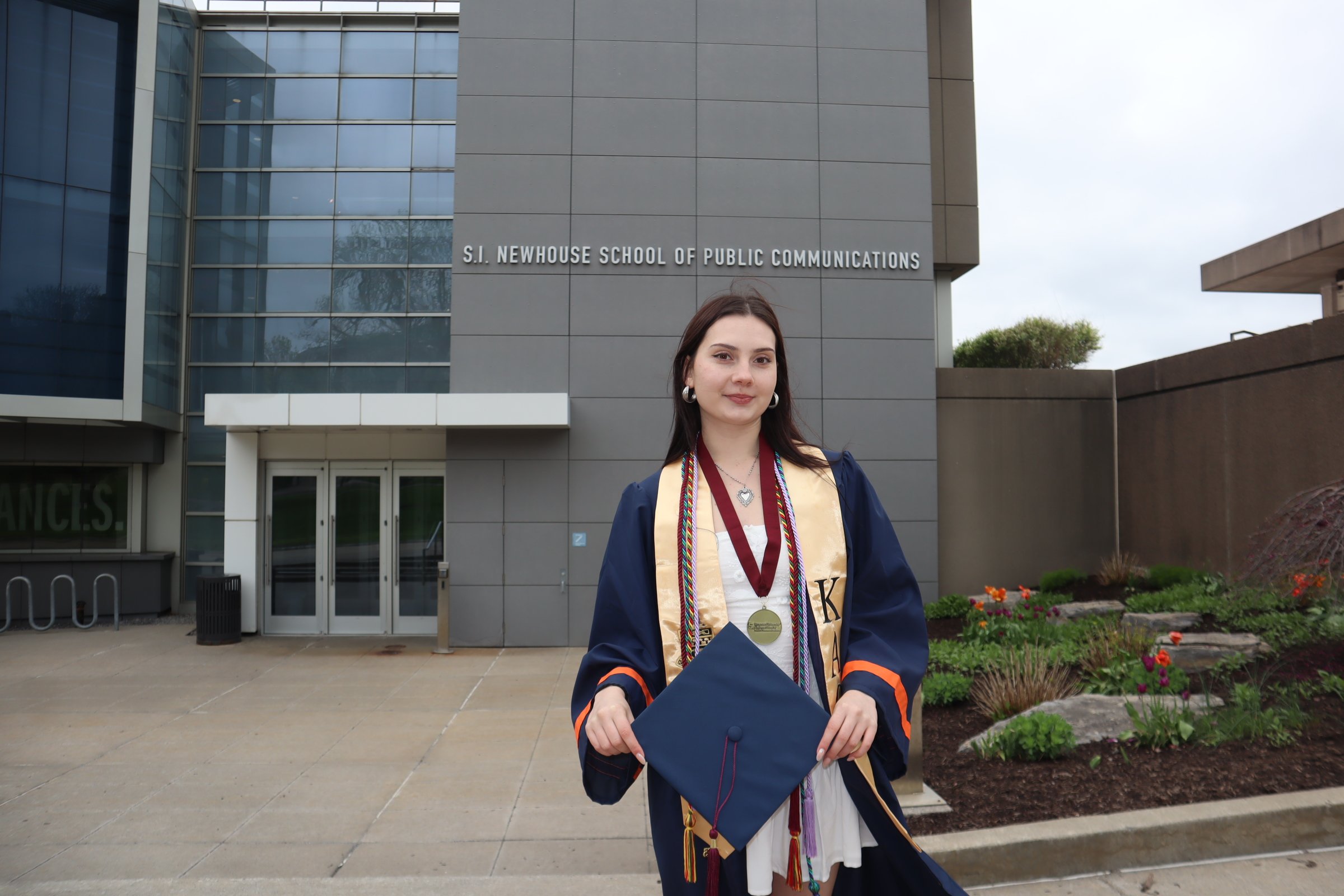 A young woman in graduation gown and cap standing outside the S.I. Newhouse School of Public Communications building holding her diploma cover and wearing medals and cords, celebrating her graduation.
