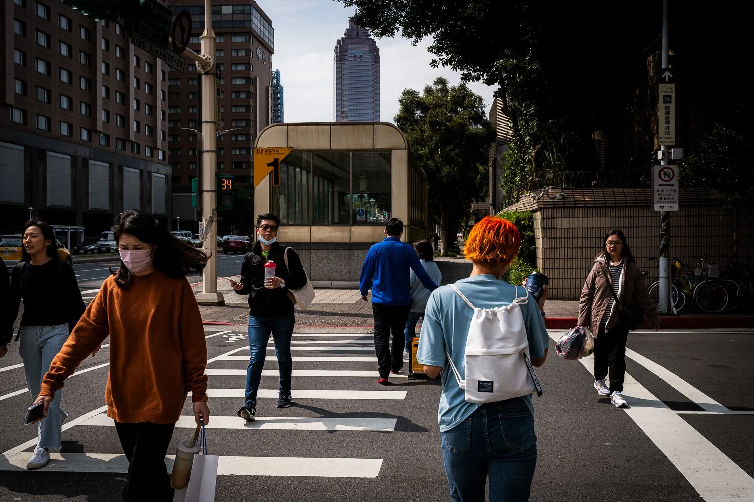 People crossing a city street at a crosswalk in front of tall buildings, some wearing masks, with trees and a bus stop in the background.