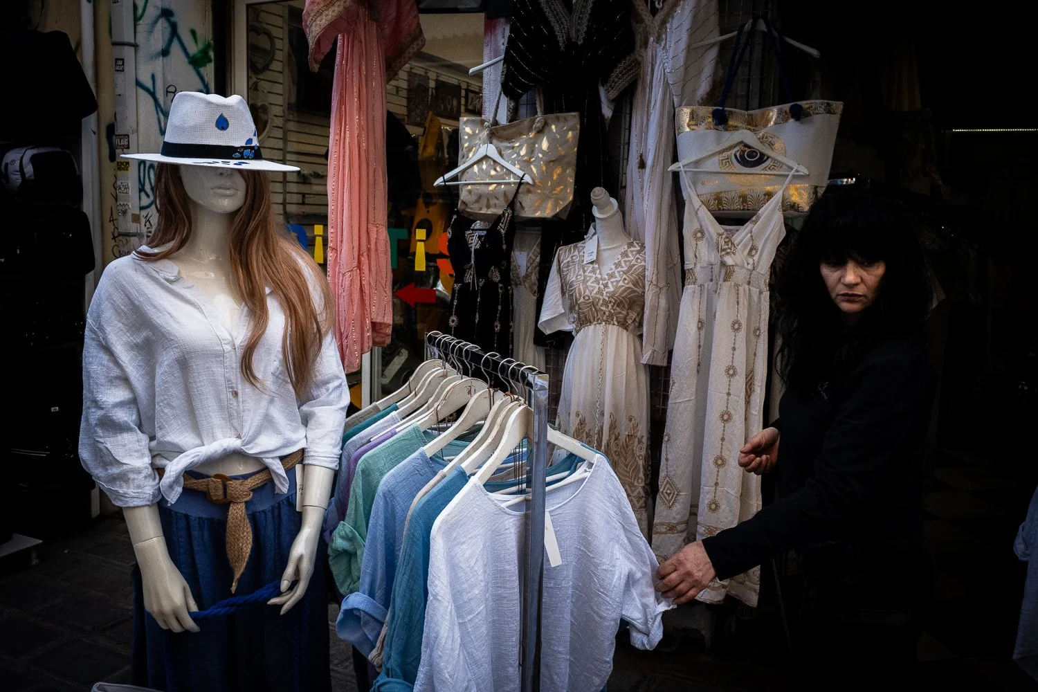 A mannequin dressed in a white button-up shirt, blue pants, a brown belt, and a wide-brimmed hat, standing in front of a clothing rack with colorful garments. An older woman with dark, shaggy hair and a black jacket is browsing clothes behind the rac