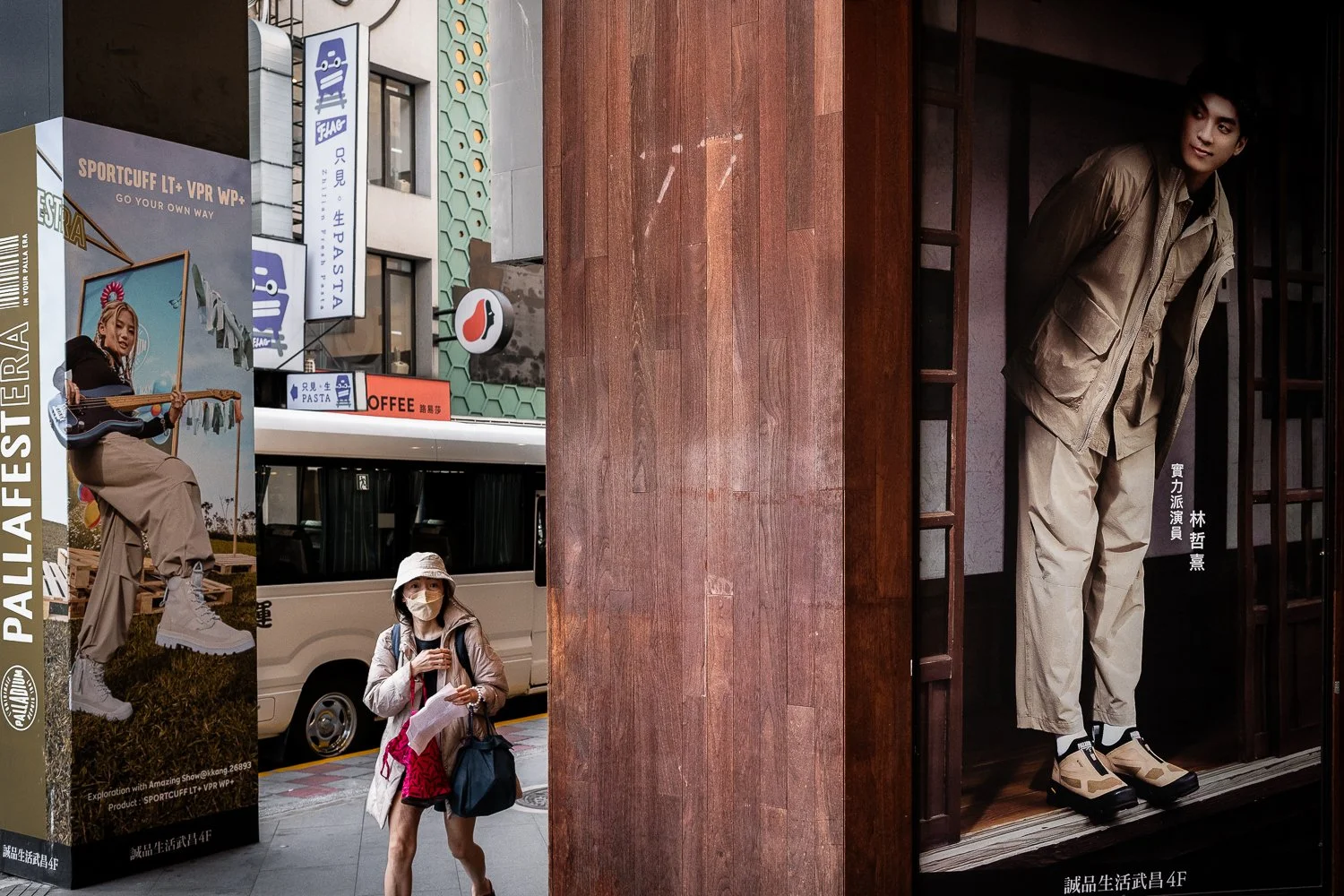 Street scene with a woman wearing a mask and carrying a bag walking past a large poster of a man in beige outdoor clothing inside a wooden frame, with various storefront signs in Asian characters in the background.