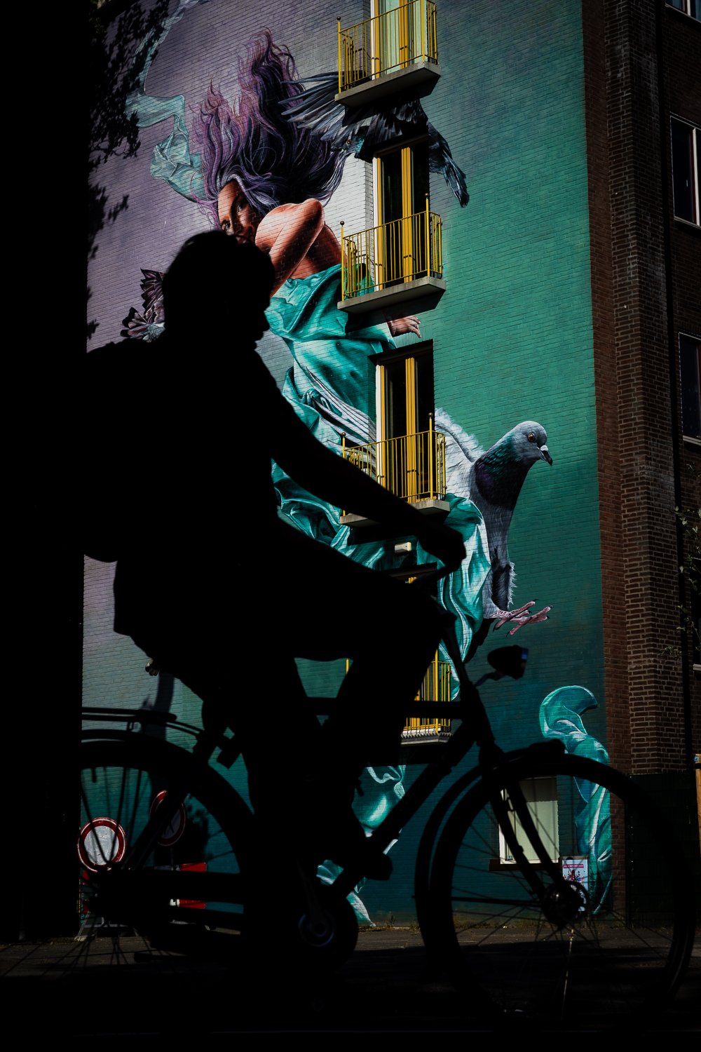 Silhouette of a person riding a bicycle in front of a colorful mural on a building wall. The mural depicts a woman with long hair and a pigeon.