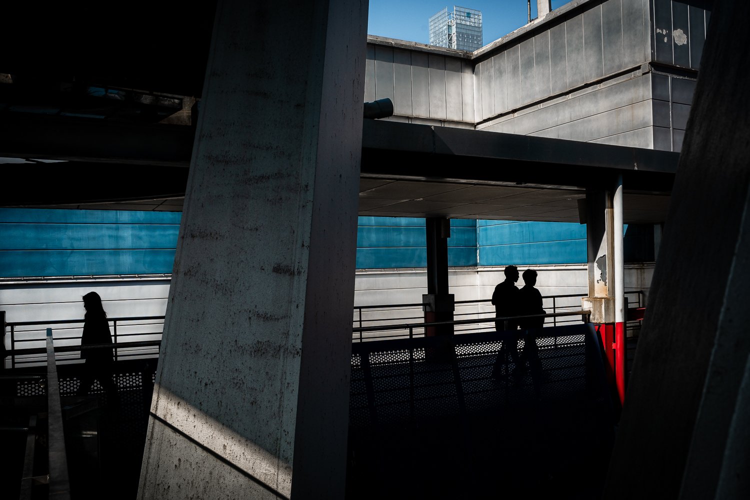 Silhouettes of three people on a higher level of an industrial or urban building, with large concrete and metal structures, in bright sunlight.