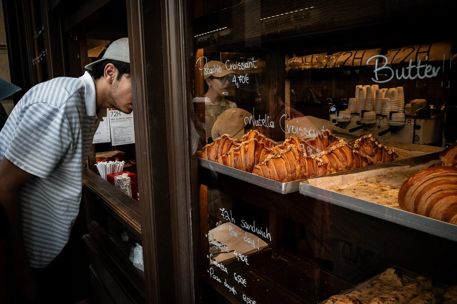 A man examining baked goods at a bakery in Athens with croissants and baked pastries inside a display case behind a glass window. The man's face is hidden behind a wooden post of the bakery