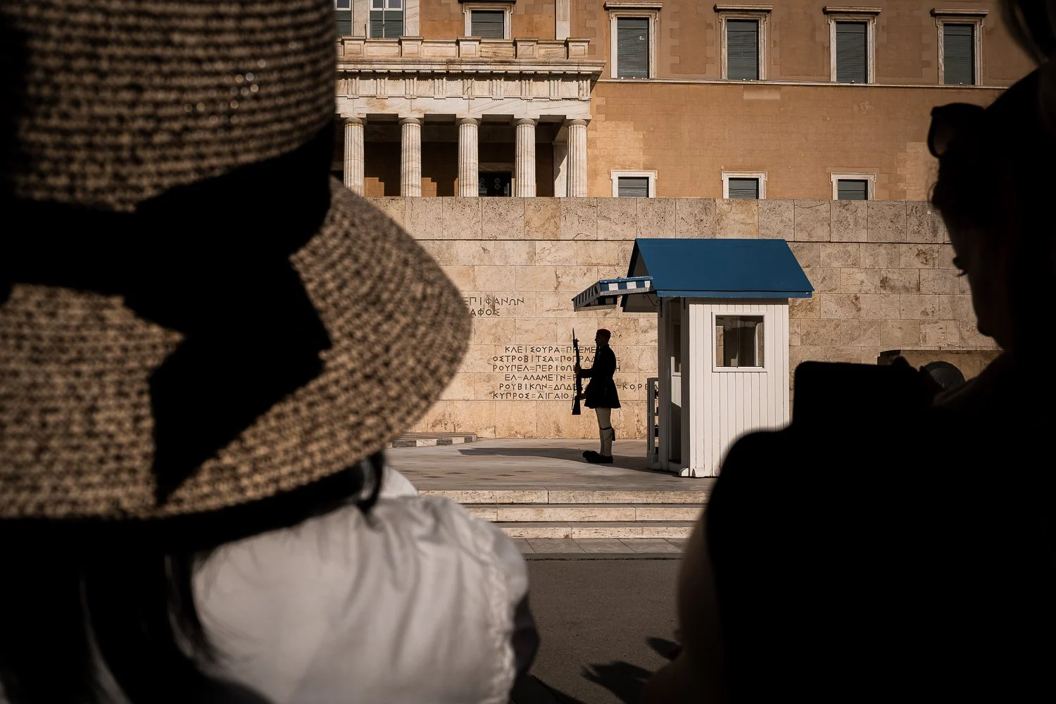 A small white and blue guard booth in front of the parliament of Greece in Athens, with a guard next to it and a that woman stands in the foreground