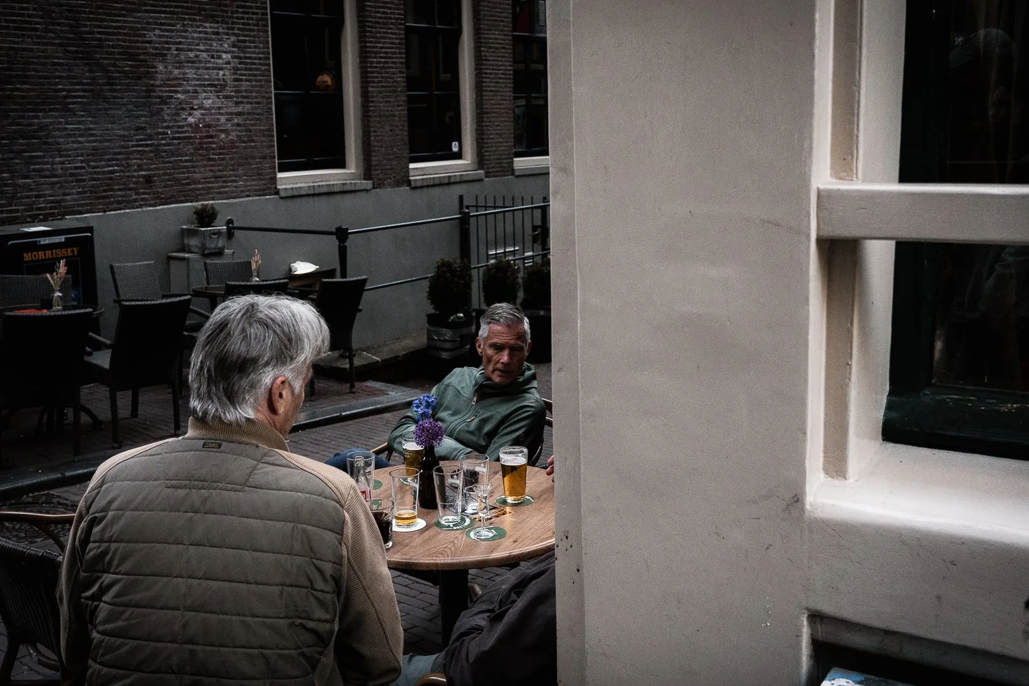Two men sitting at a table outdoors, having drinks, viewed through a partial wall with a window, in an urban setting with empty chairs and potted plants in the background.