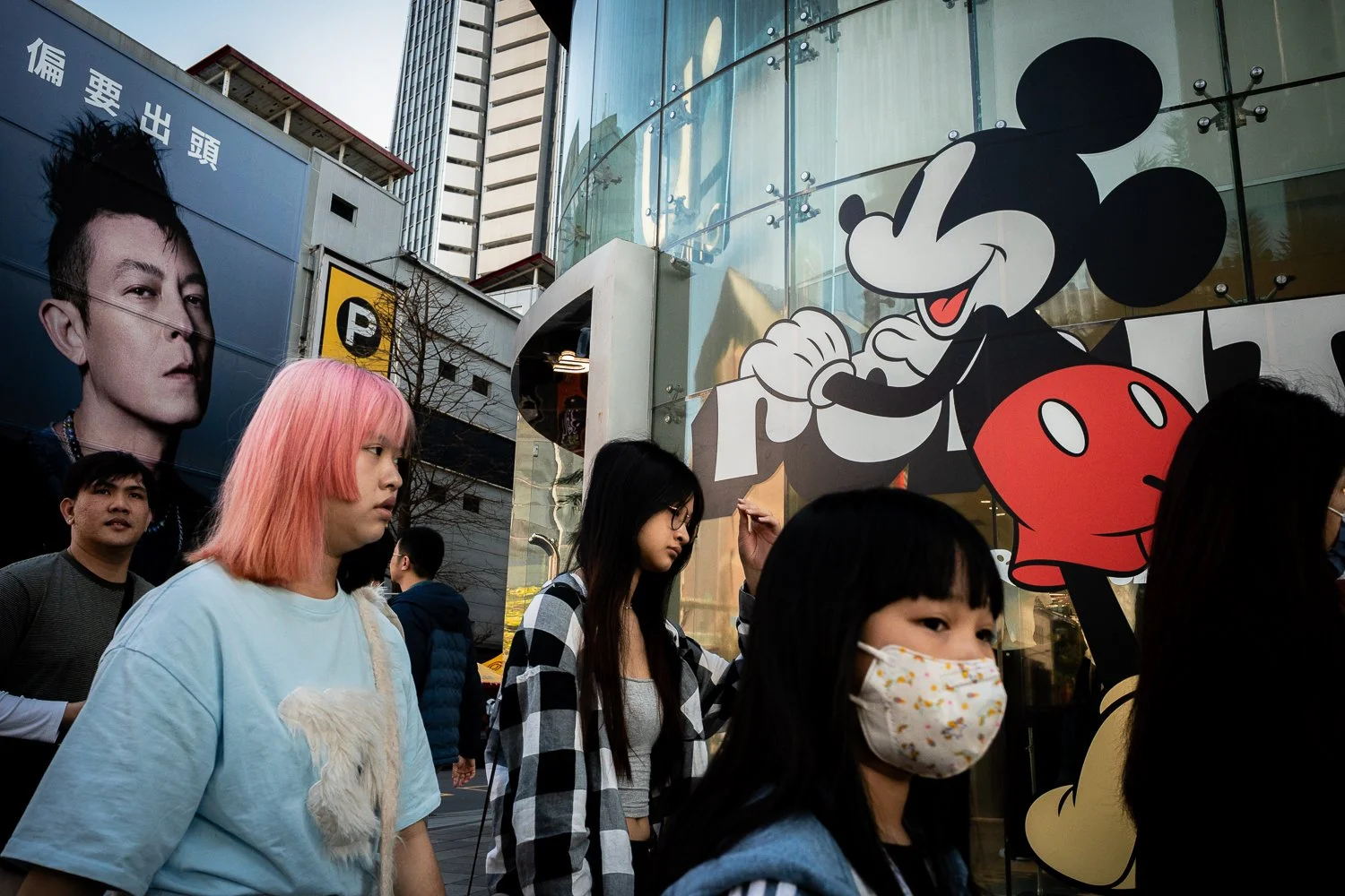 People walking past a shop with a large Mickey Mouse graphic on the window, with several pedestrians including a girl with pink hair and a face mask.