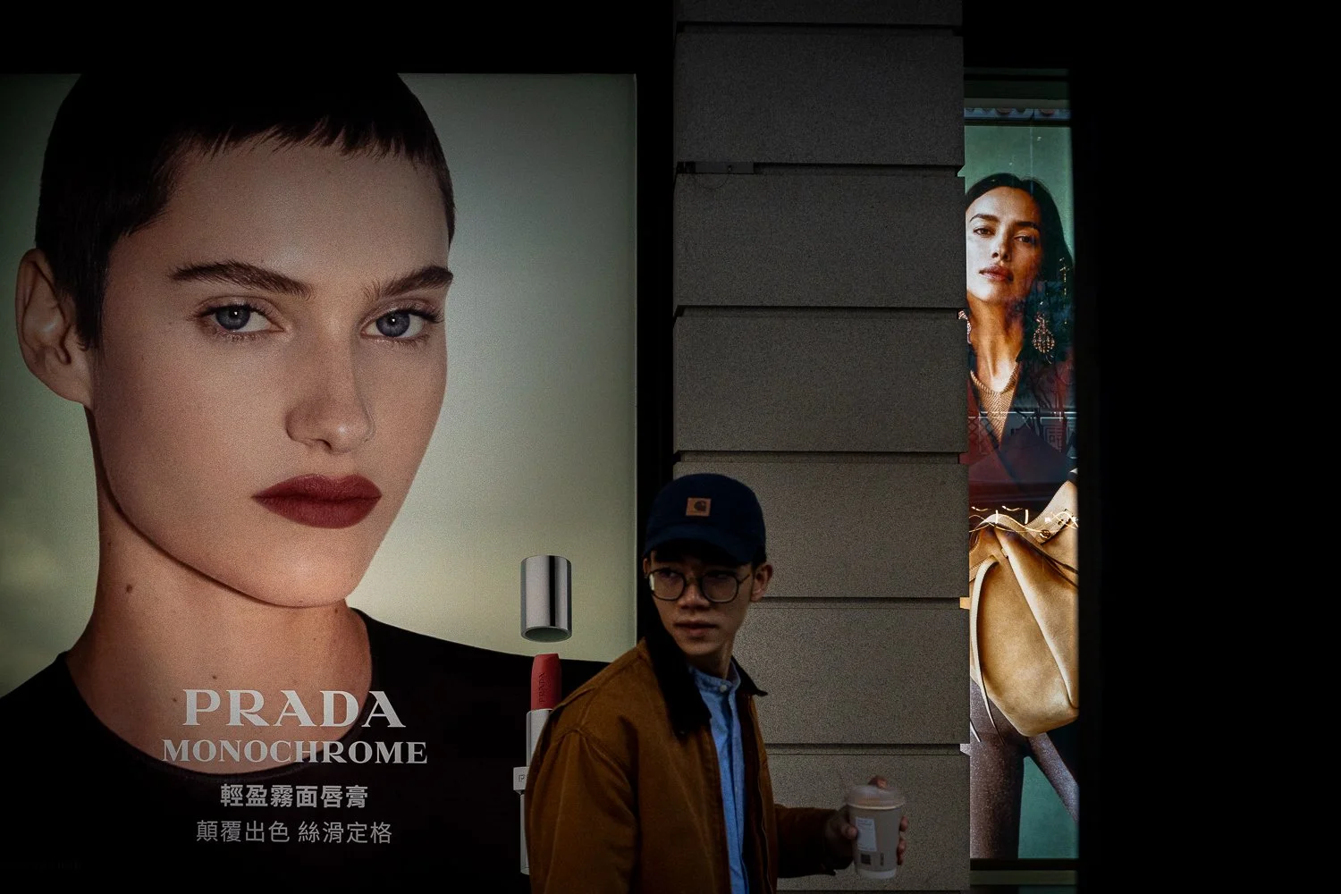 A young man wearing glasses and a baseball cap holding a coffee cup walks past a Prada Monochrome advertisement featuring a close-up of a woman's face with red lips and short hair, with another advertisement of a woman in jewelry visible behind a win