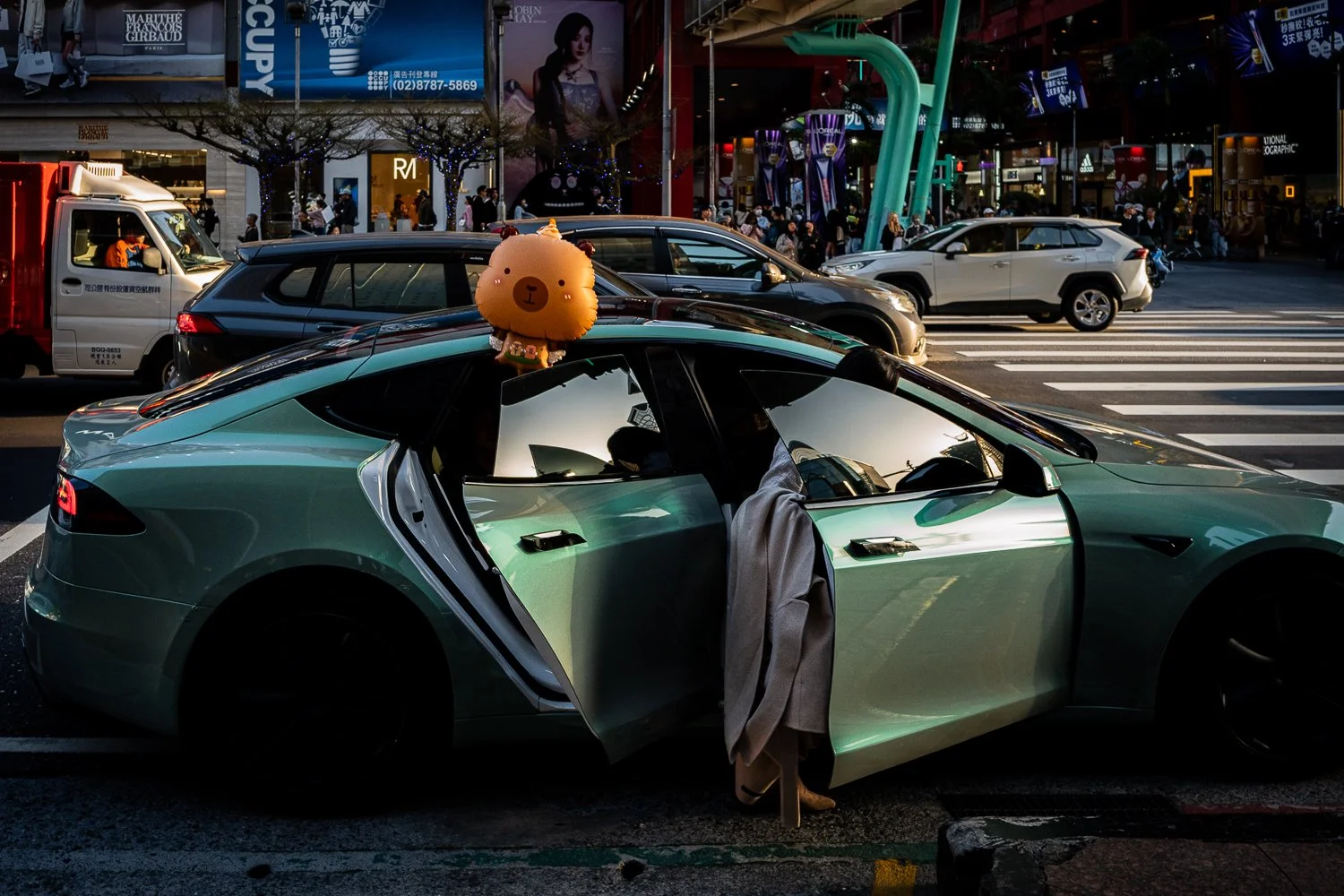 Green car parked at an intersection with a bear-shaped balloon floating out of the car window and a person entering through the open driver's side door. The background shows busy city traffic and illuminated advertisements.
