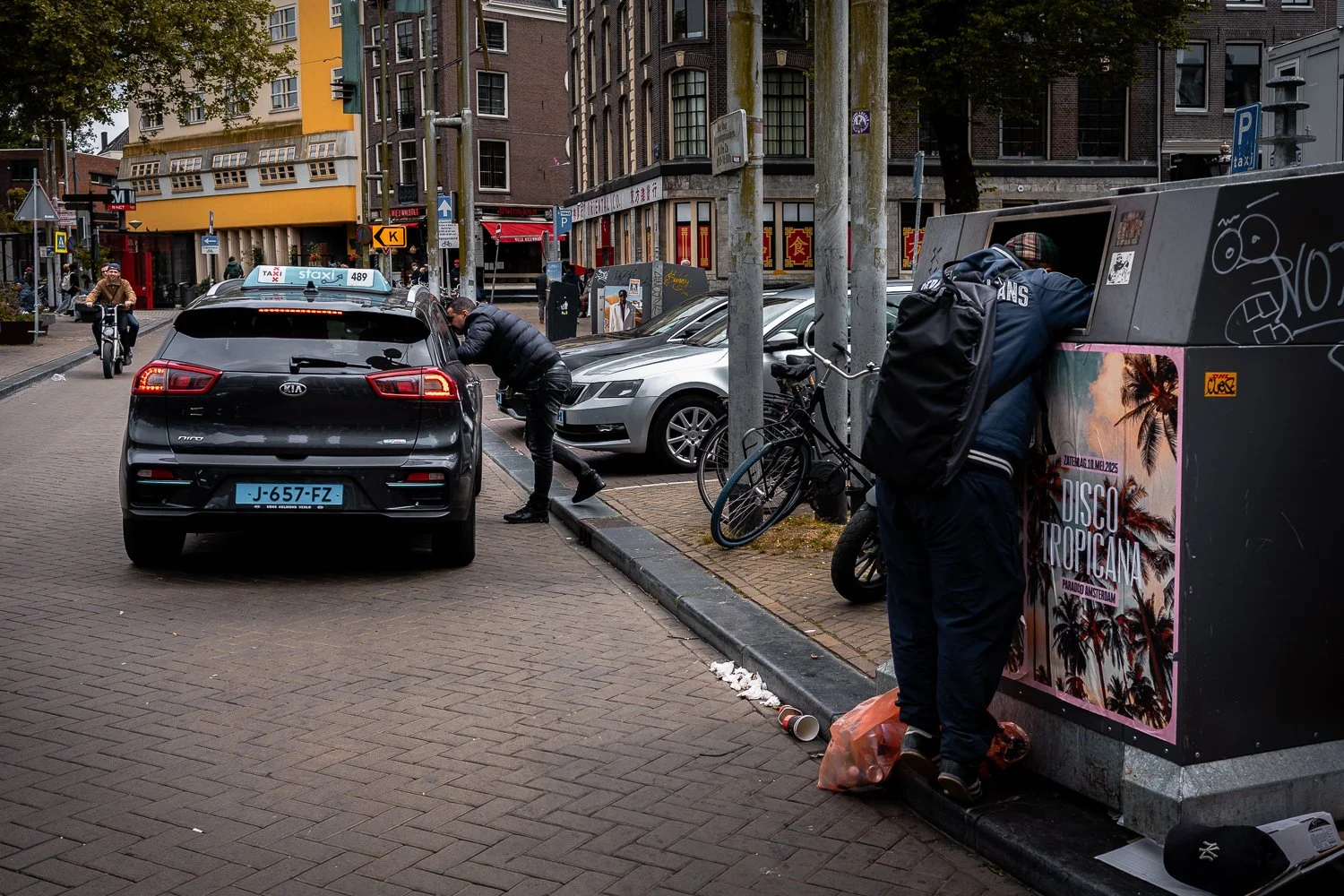 People at a street-side parking area, with one person leaning into a parking meter and another looking into a trash bin. Several parked cars and bicycles are visible, along with residential and commercial buildings in the background.