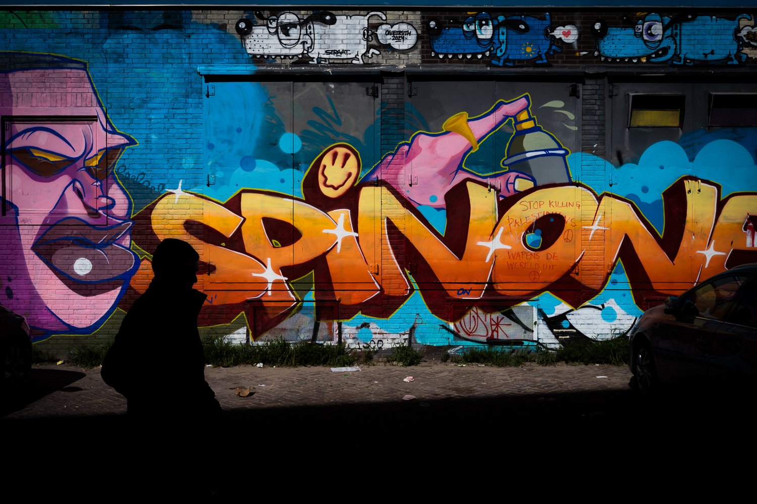 Colorful graffiti art on brick wall featuring a large word, animated characters, and a silhouette of a person walking in front.