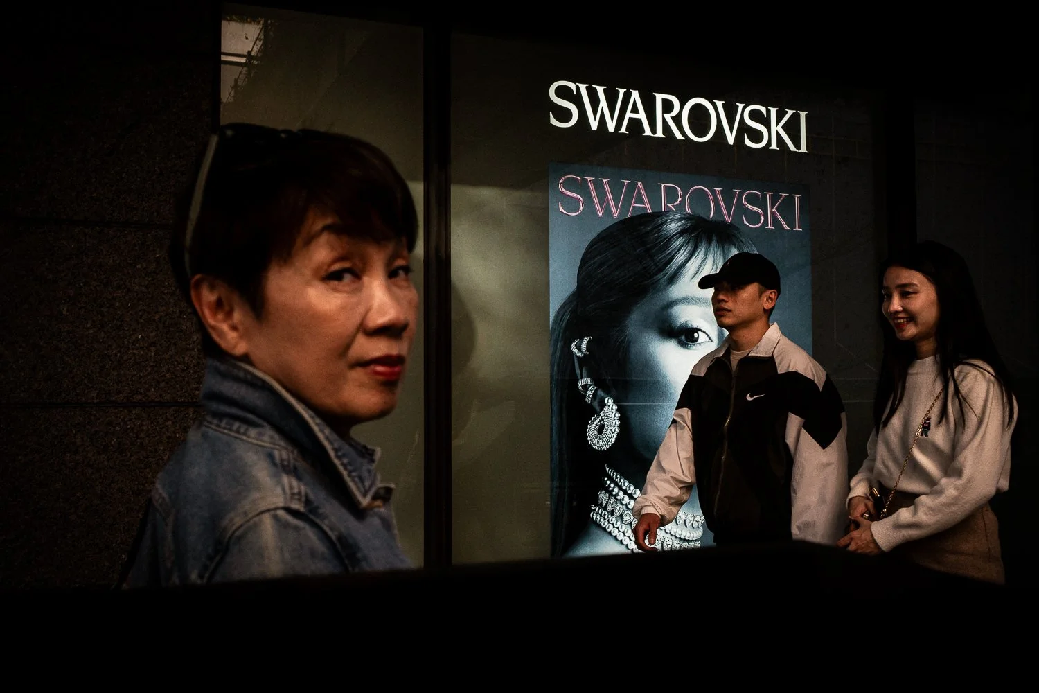 Three people standing in front of a Swarovski store window display, with one woman looking at the camera and a man and woman talking and smiling in the background.