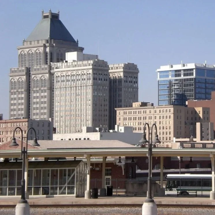 City skyline with tall buildings, including one with a distinctive sloped roof, and a train station platform with street lamps in the foreground.