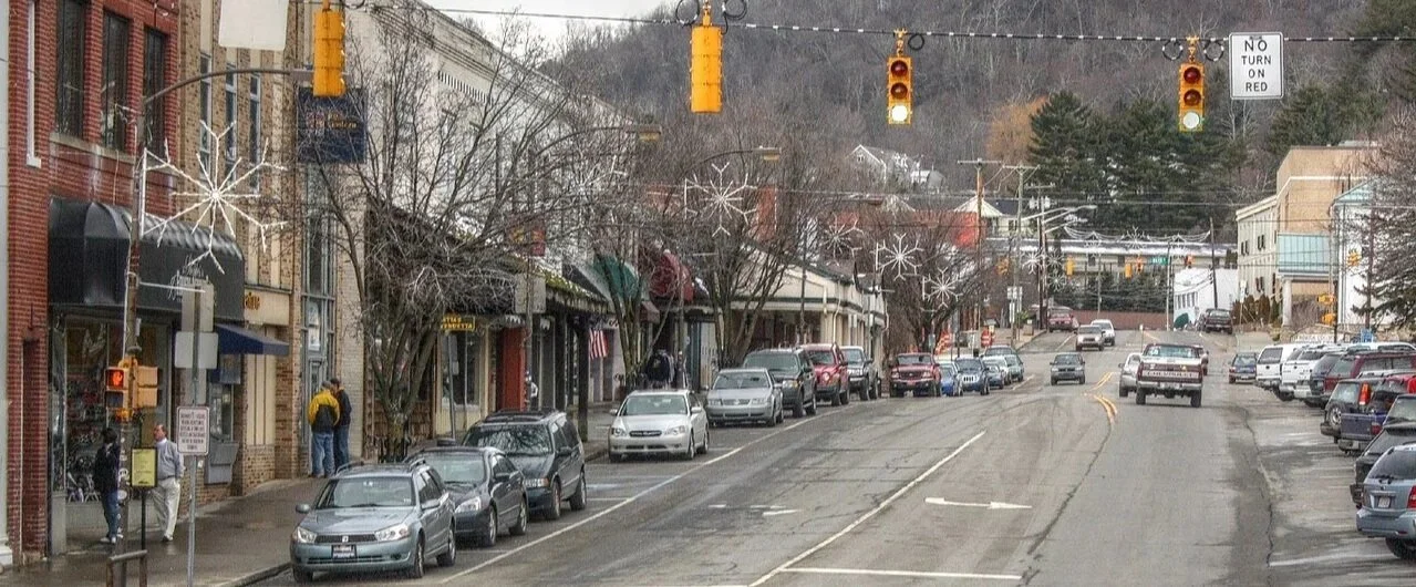 Downtown street scene with parked cars, storefronts, leafless trees, traffic lights, and a "No Turn on Red" sign, under an overcast sky.