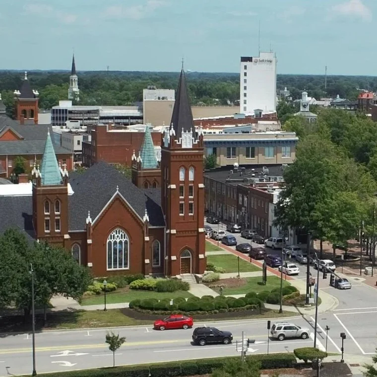 A cityscape with a red brick church featuring gothic architecture and tall spires in the foreground, surrounded by trees and parked cars, with commercial buildings and a white high-rise in the background under a partly cloudy sky.
