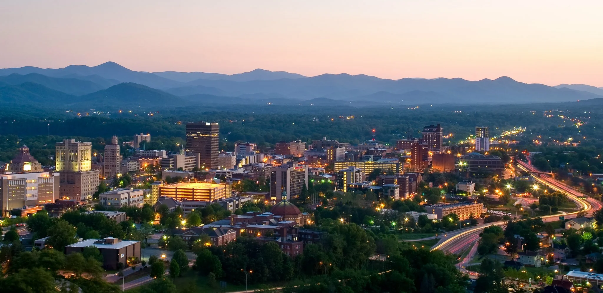 Cityscape at dusk with illuminated buildings, winding roads, and mountain range in the background