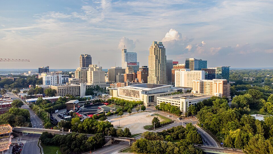 A city skyline with tall buildings, some with glass windows, surrounded by trees and roads under a partly cloudy sky.