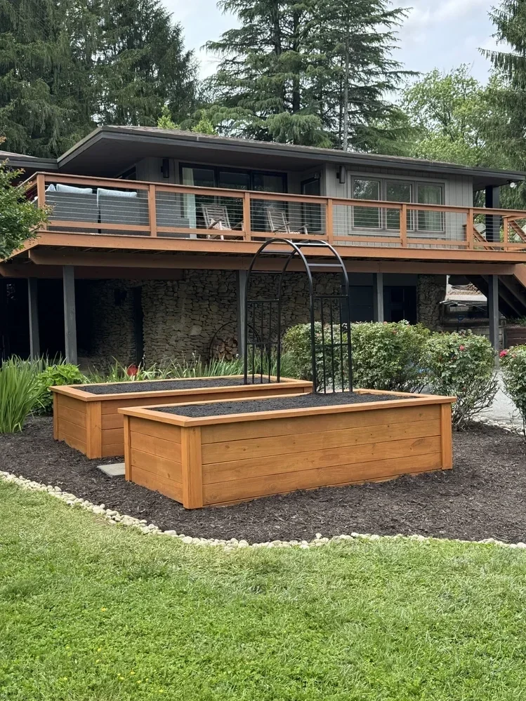 Raised wooden garden beds with soil in front of a house with a balcony, surrounded by greenery.