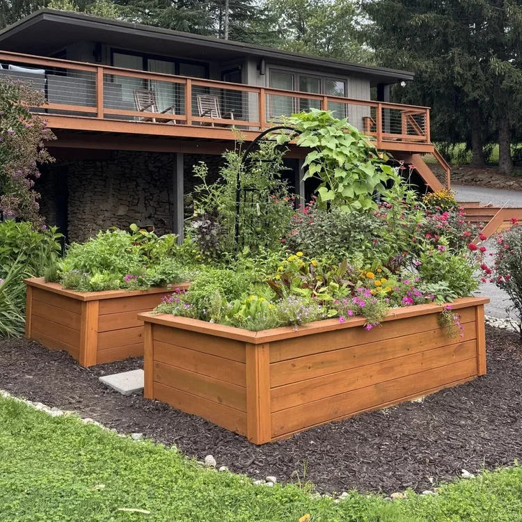 Raised wooden garden beds filled with blooming flowers and vegetables in front of a modern house with a balcony and outdoor seating area.
