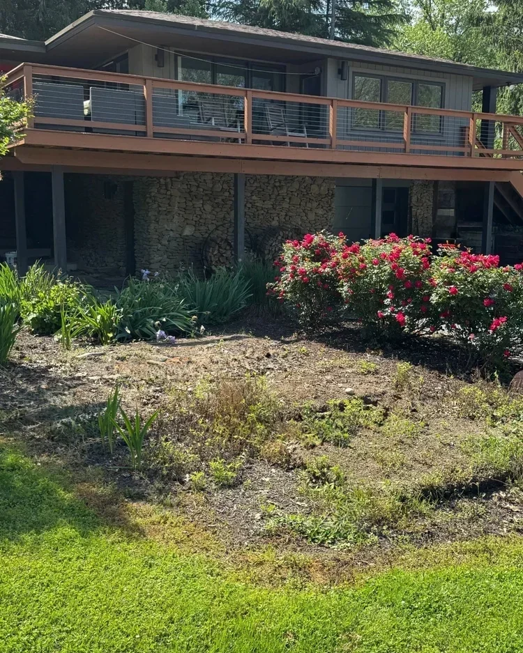 A house with a large wooden deck, stone foundation, and large glass windows. There are shrubbery and flowering plants in the garden in front of the house.