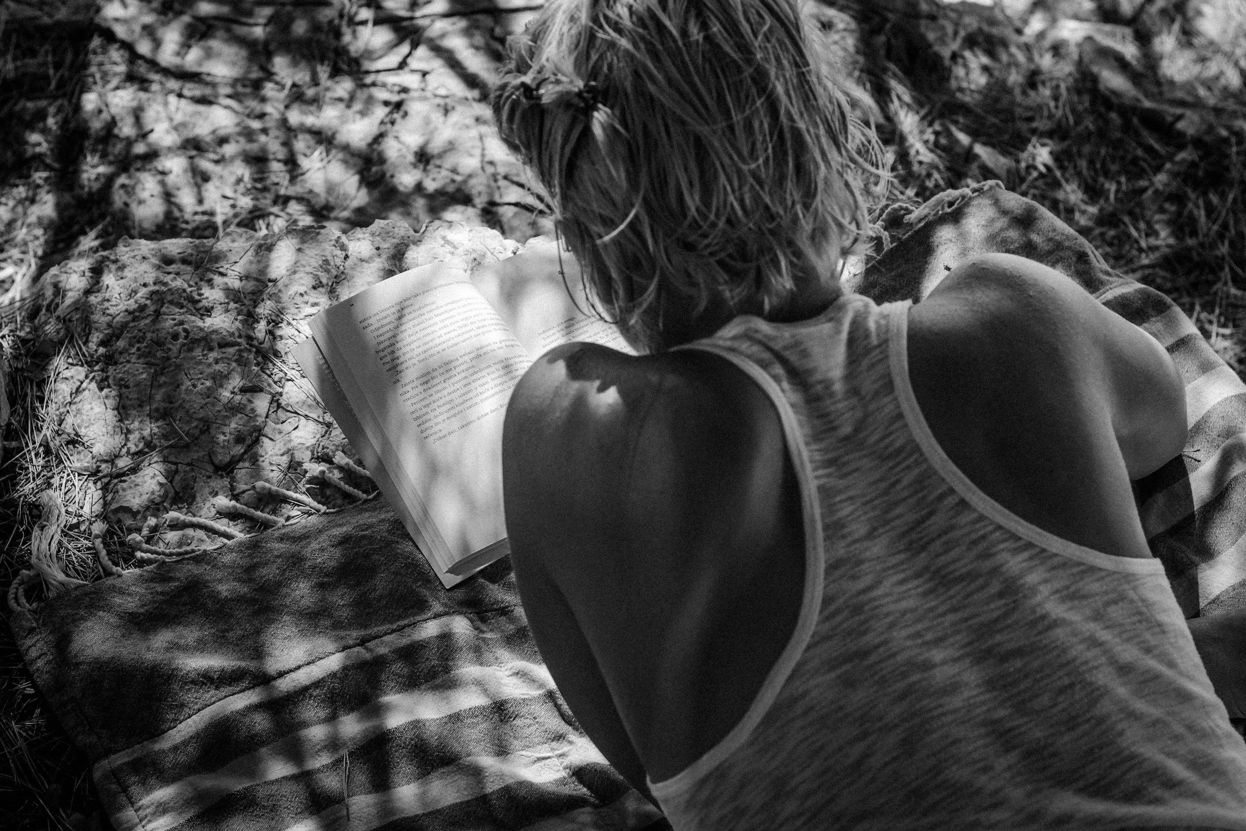 woman with short hair, wearing a white tank, laying on sarong, reading a book.
