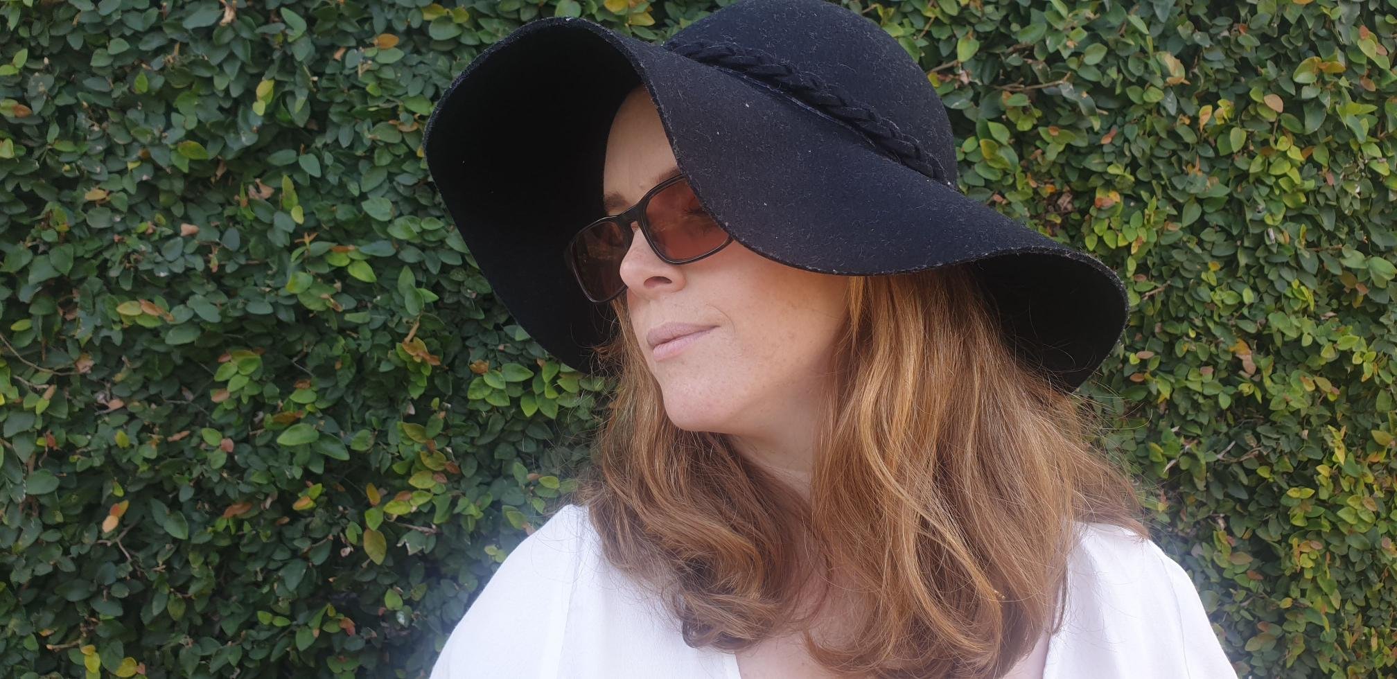 headshot of Jess. She has long red hair and is wearing a navy blue felt hat, sunglasses and a white top.