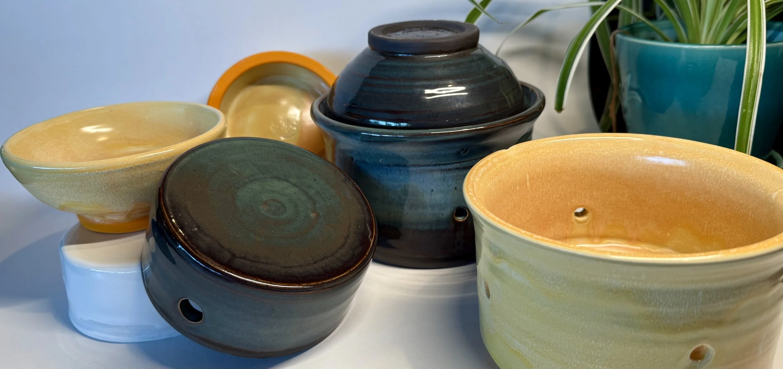 Collection of handmade ceramic tofu-making kits in various colors including yellow, and green and blue, with some having holes or lids, arranged on a white surface near a green potted plant.